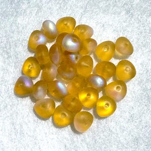 May include: A collection of small, translucent yellow beads with an iridescent finish. Each bead features a rounded, irregular shape and a central hole for threading. The surface of the beads displays a soft, pearlescent sheen that reflects light.