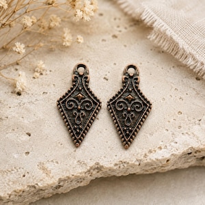 May include: Two identical diamond-shaped metal charms with an antique copper finish. Each charm features an intricate embossed scrollwork pattern, a beaded border, and a small circular loop at the top for attachment to jewellery or craft projects.