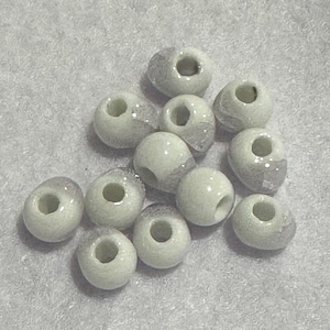 May include: A collection of off-white, round beads with a hole through the center. Some beads have a silver-colored coating on one side. The beads are scattered on a light gray surface. These beads are suitable for jewelry making.