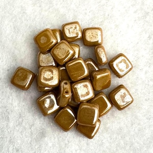 May include: A collection of small, square-shaped beads with a smooth, glossy finish in a warm, metallic bronze color. Each bead features rounded corners and a central hole, displaying a reflective surface that catches light across the pile.