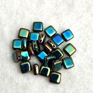 May include: A collection of small, square-shaped beads with a metallic, iridescent finish. The beads display a vibrant spectrum of colors, shifting between deep blue, teal, and hints of purple and gold, each featuring two small holes for threading.