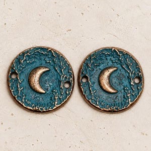 Puede incluir: Dos dijes metálicos circulares con una media luna en relieve de color bronce en el centro. La superficie circundante tiene un acabado de pátina azul verdoso texturizado y desgastado. Cada dije incluye dos pequeños orificios laterales.