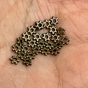 May include: A collection of small, bronze-coloured metal bead caps, each shaped like a flower with multiple petals. The bead caps have a central hole for stringing and are scattered across a neutral background.