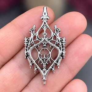 May include: A silver-toned metal pendant featuring an intricate, openwork design with pointed architectural elements and symmetrical filigree patterns. The piece has a central vertical structure with sharp, decorative finials at the top and bottom.