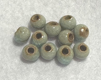 May include: A collection of pale green ceramic beads with a glossy finish. Each bead has a central hole and a slightly irregular shape. The beads are scattered on a light-colored surface, suitable for jewelry making or crafts.
