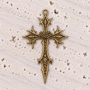 May include: An antique brass-toned metal cross pendant featuring intricate filigree detailing, pointed flared ends, and a central rose motif. The cross has a small circular loop at the top for attachment and displays a textured, weathered finish.