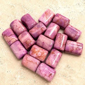 May include: A collection of fifteen polished, cylindrical beads made of pink and purple stone with natural veining and marbled patterns. Each bead features a smooth, glossy finish and a uniform shape, arranged in a loose, heart-like formation.