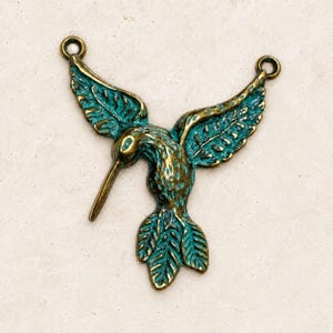May include: A hummingbird-shaped metal pendant featuring a weathered bronze and teal patina finish. The bird is depicted in flight with wings spread wide, a long pointed beak, and two small circular loops at the top of each wing for attachment.