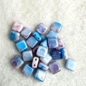 May include: A collection of small, square-shaped beads featuring a smooth, glossy finish. Each bead displays a marbled pattern in shades of sky blue, deep indigo, and soft white, with some pieces showing subtle hints of pinkish-purple hues.