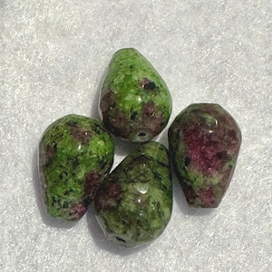 May include: Four green and maroon gemstone beads with a teardrop shape. The beads have a polished surface and are arranged on a white surface. The beads are likely for jewelry making.
