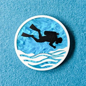 May include: A white circle with a blue background and a black silhouette of a scuba diver with a tank on their back. The diver is surrounded by white waves.