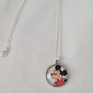 May include: A silver necklace with a circular pendant depicting Mickey Mouse in a red shirt. The pendant hangs from a fine chain with a clasp. The cartoon character is in a classic pose.