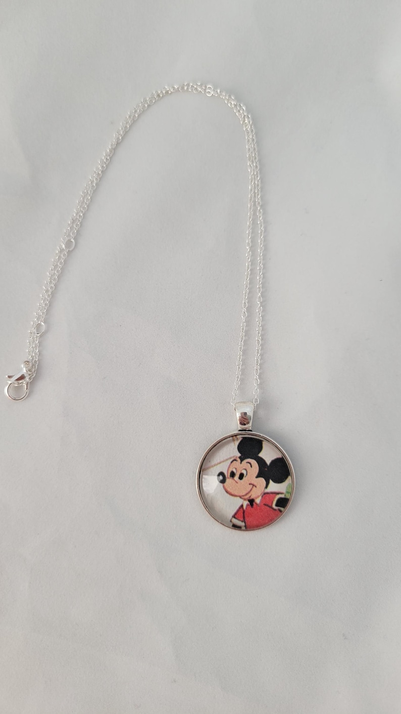May include: A silver necklace with a circular pendant depicting Mickey Mouse in a red shirt. The pendant hangs from a fine chain with a clasp. The cartoon character is in a classic pose.
