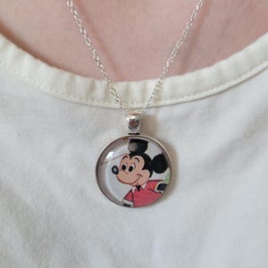 May include: Silver necklace with a circular pendant displaying a cartoon image of Mickey Mouse. Mickey is depicted in his iconic red jacket and black ears. The necklace is set against a white backdrop.
