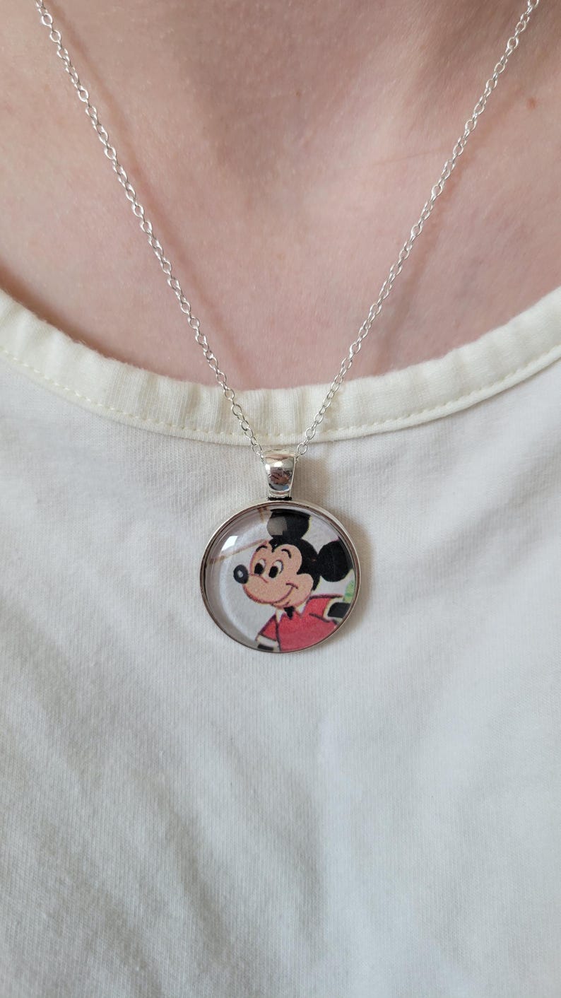 May include: Silver necklace with a circular pendant displaying a cartoon image of Mickey Mouse. Mickey is depicted in his iconic red jacket and black ears. The necklace is set against a white backdrop.