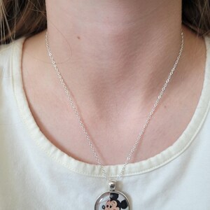 May include: A silver necklace with a circular pendant displaying a cartoon image of Mickey Mouse. The pendant has a clear cover. The necklace is worn against a white t-shirt.