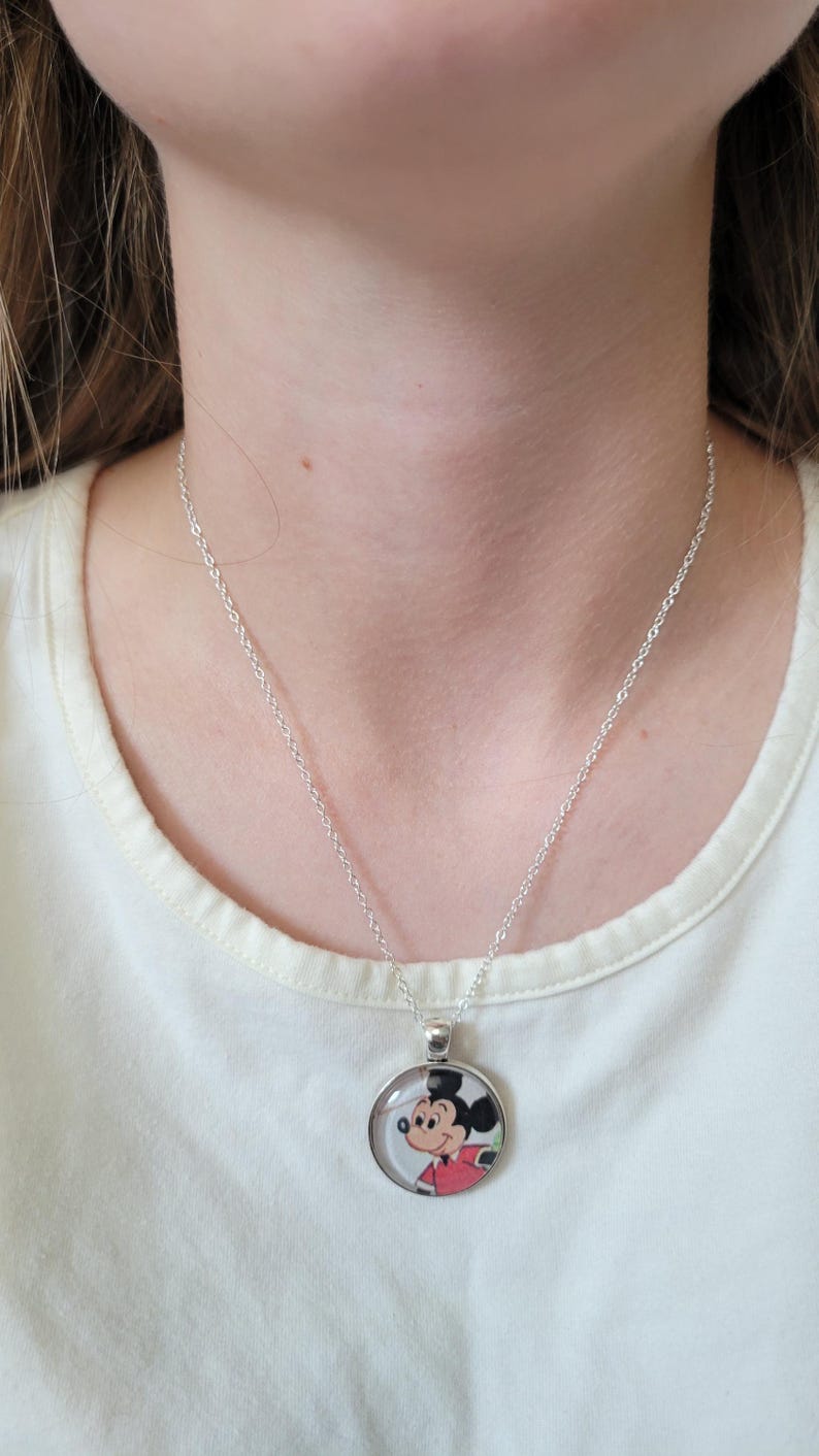 May include: A silver necklace with a circular pendant displaying a cartoon image of Mickey Mouse. The pendant has a clear cover. The necklace is worn against a white t-shirt.