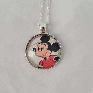 May include: A silver necklace with a circular pendant displaying a cartoon image of Mickey Mouse. Mickey is depicted in a red jacket and black ears. The pendant hangs from a fine silver chain. The background is a plain white surface.