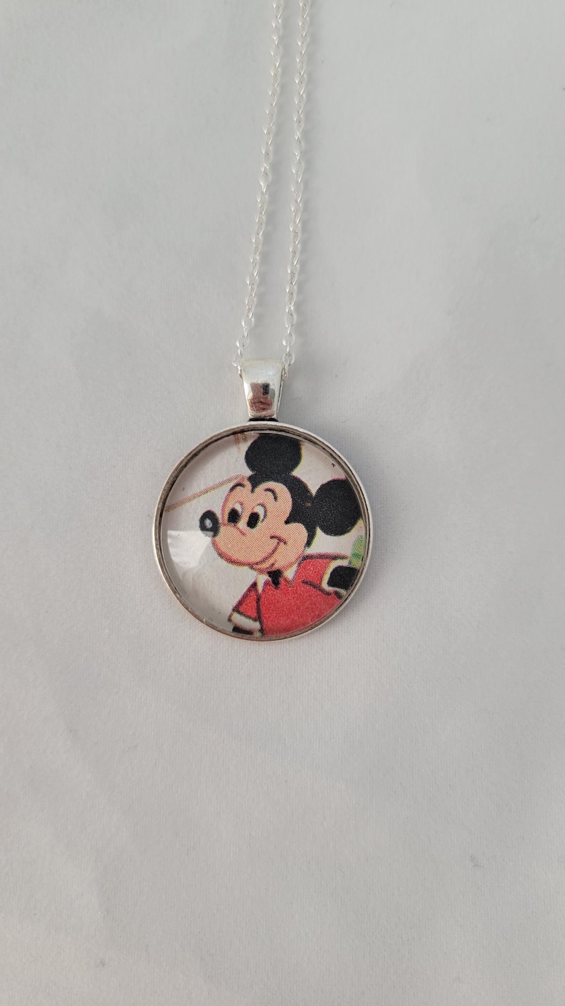 May include: A silver necklace with a circular pendant displaying a cartoon image of Mickey Mouse. Mickey is depicted in a red jacket and black ears. The pendant hangs from a fine silver chain. The background is a plain white surface.
