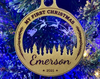 Op de afbeelding: Een gouden houten kerstversiering met een transparant venster dat een silhouet laat zien van een slee die door rendieren wordt getrokken. De versiering is versierd met een bruine krans en de tekst "My First Christmas Emerson * 2021 *"
