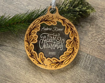 Op de afbeelding: Een ronde, houten kerstbal met een transparant glazen midden. De bal is versierd met een gouden bloemenkrans en de tekst "Aubree Renee First Christmas 2022".