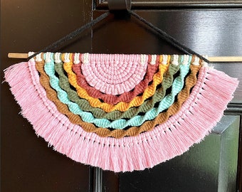 Puede incluir: Un tapiz de pared de crochet rosa, marrón, naranja, amarillo, verde y blanco con flecos. El tapiz tiene forma de semicírculo.