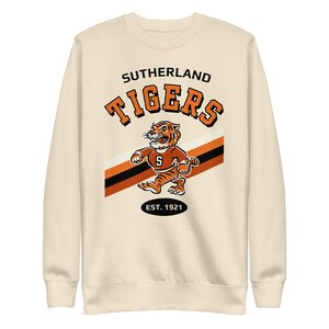 May include: A cream colored sweatshirt with a vintage-style graphic of a tiger mascot. The graphic includes the text "Sutherland Tigers" and "Est. 1921".