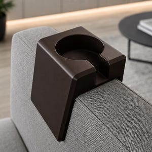 May include: A dark brown, saddle-shaped cup holder designed to drape over the arm of a sofa. The accessory features a circular cutout for a beverage container and a rectangular notch on the front edge, constructed from a solid, smooth material.