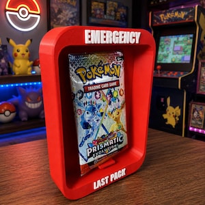 May include: A bright red rectangular display frame with rounded corners featuring the white text EMERGENCY at the top and LAST PACK at the bottom. A sealed Pokémon Scarlet and Violet Prismatic Evolutions trading card game booster pack is centered.
