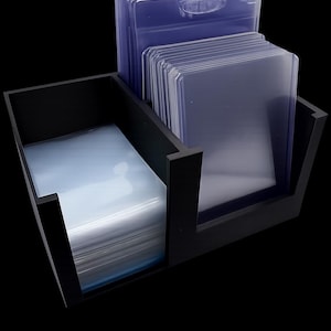 May include: A black, two-compartment card holder with stacks of clear, protective card sleeves. The sleeves are rectangular and appear to be made of a transparent plastic material. The holder is made of a dark, possibly painted, wood or composite material.