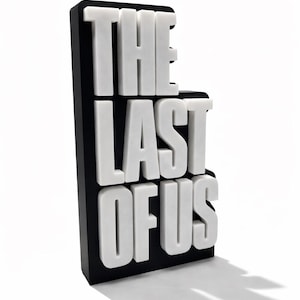 May include: A 3D sign featuring the text "THE LAST OF US" in white, block-style letters against a black background. The sign casts a shadow on the white surface below.