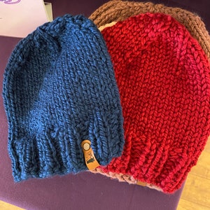 May include: Three knitted hats in blue, red, and brown. The blue hat has a leather tag with a design on it.