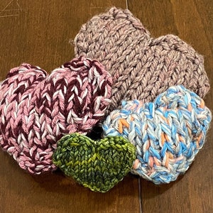 May include: A collection of five knitted heart-shaped items in various colors. The hearts are in shades of pink, purple, blue, green, and a neutral tone. The knitted texture is visible, with each heart displaying a unique color combination.