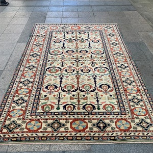 Puede incluir: Una alfombra rectangular de color crema con un patrón intrincado. La alfombra presenta un diseño central con motivos florales y geométricos repetidos en tonos de rojo, azul, verde y negro. Un borde oscuro enmarca el diseño.