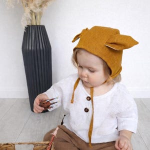 May include: A baby wearing a mustard-colored hat with ear-like flaps, a white button-down shirt, and brown shorts. The baby is holding a toy and sitting near a wicker basket with toy animals.