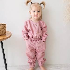 Puede incluir: Un conjunto de dos piezas en color rosa empolvado que consta de una parte superior de manga larga con cuello redondo y tres botones frontales, junto con pantalones tipo jogger a juego con cintura el&aacute;stica, pu&ntilde;os y bolsillos laterales.