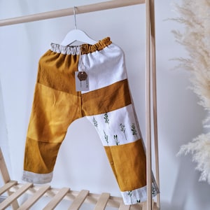 May include: Mustard yellow and white patchwork pants with a floral print on the lower leg. The pants are hanging on a wooden rack. The pants have an elastic waistband and a tag attached. The pants are made of a soft, lightweight fabric.