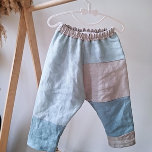 May include: Patchwork baby trousers in shades of blue, tan, and beige, hanging on a white hanger. The trousers have an elasticated waistband and a relaxed fit. The fabric appears to be linen or a similar lightweight material. The trousers are displayed on a wooden clothing rack.
