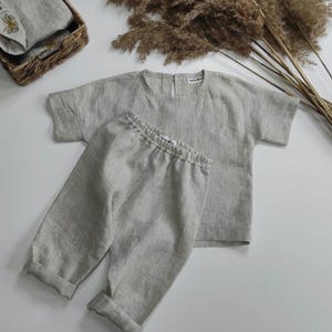 May include: A light gray linen two-piece set consisting of a short-sleeved top with a round neckline and a button closure at the back, and matching trousers with an elastic waistband and cuffed hems. Both pieces feature a small white brand label.