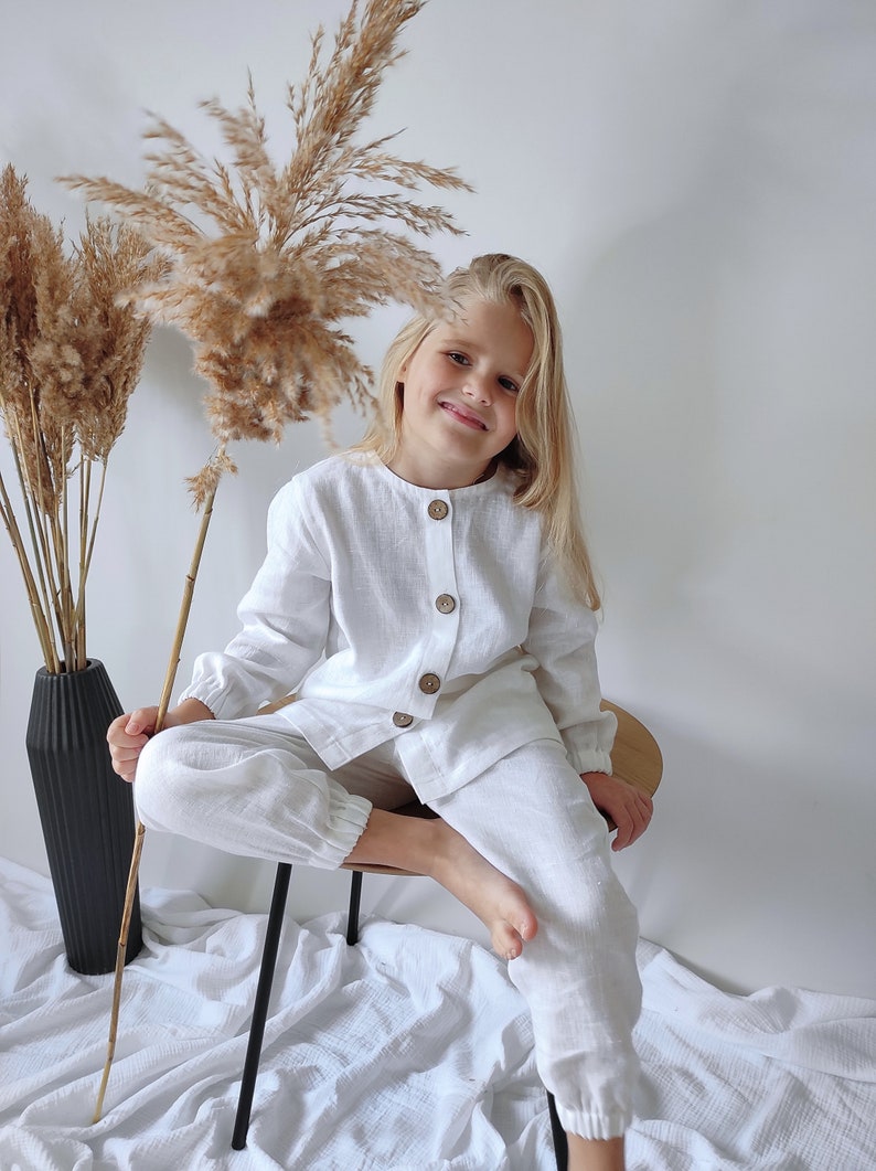 Puede incluir: Un conjunto de camisa blanca de manga larga con botones y pantal&oacute;n a juego estilo jogger. La camisa presenta un cuello redondo y cuatro botones de madera marr&oacute;n en la parte delantera. El pantal&oacute;n tiene pu&ntilde;os el&aacute;sticos en los tobillos.