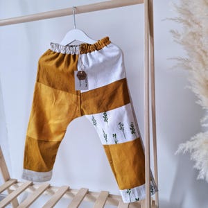 May include: Pants featuring a color-blocked design in mustard yellow and white. The white sections are decorated with a small green botanical print. The garment has an elasticized waistband and is suspended from a white plastic hanger.