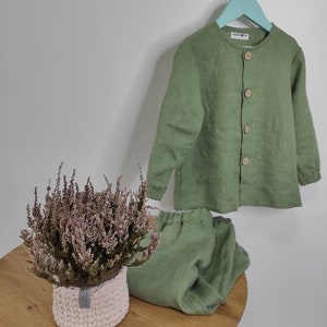 Puede incluir: Un conjunto de camisa de manga larga color verde salvia con botones y pantal&oacute;n a juego con cintura el&aacute;stica. La camisa presenta cuello redondo, cuatro botones de madera clara y pu&ntilde;os el&aacute;sticos. Se observa una etiqueta blanca con el texto "byanka & lia".