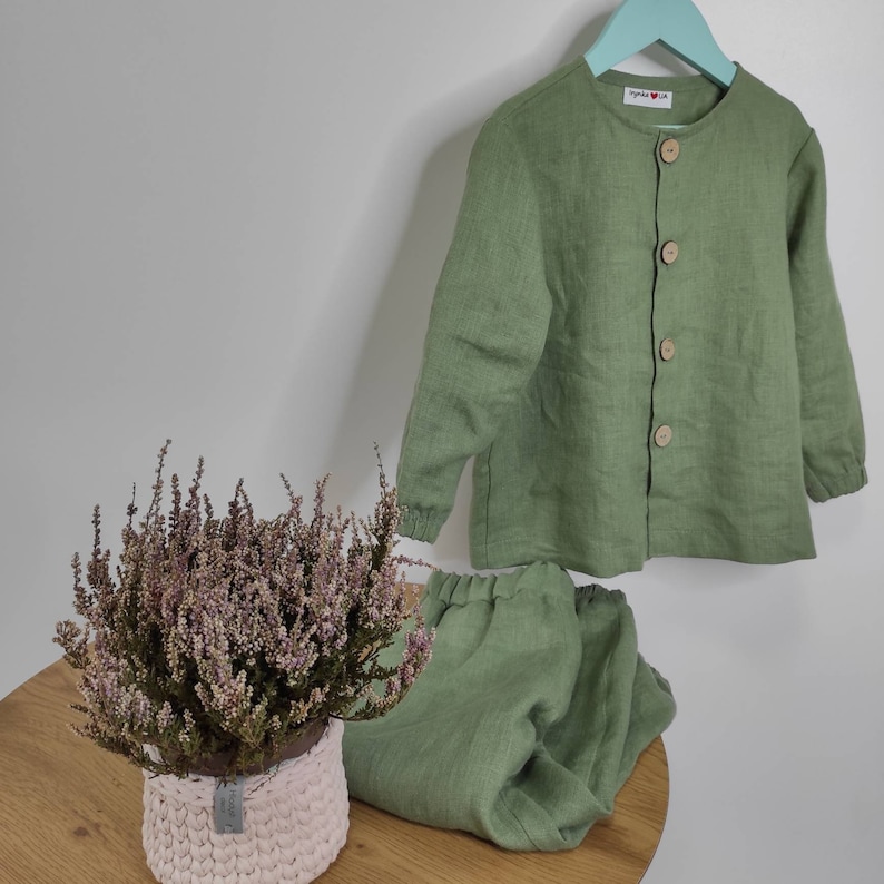 Puede incluir: Un conjunto de camisa de manga larga color verde salvia con botones y pantal&oacute;n a juego con cintura el&aacute;stica. La camisa presenta cuello redondo, cuatro botones de madera clara y pu&ntilde;os el&aacute;sticos. Se observa una etiqueta blanca con el texto "byanka & lia".