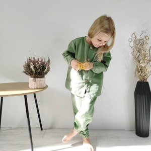 Puede incluir: Un conjunto de camisa de manga larga color verde salvia con botones y pantalones a juego con pu&ntilde;os el&aacute;sticos. La camisa presenta un cuello redondo y cuatro botones grandes de madera clara en la parte delantera. La tela tiene un tejido visible.