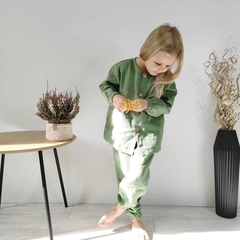 Puede incluir: Un conjunto de camisa de manga larga color verde salvia con botones y pantalones a juego con pu&ntilde;os el&aacute;sticos. La camisa presenta un cuello redondo y cuatro botones grandes de madera clara en la parte delantera. La tela tiene un tejido visible.