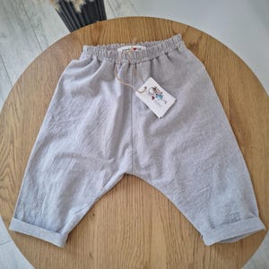 Könnte beinhalten: Hellgraue Hose mit elastischem Bund und umgeschlagenen Säumen. Die Hose hat einen tief sitzenden Schritt und ein weißes Papieretikett, das mit einer braunen Schnur befestigt ist. Das Etikett zeigt eine Illustration und den Text "mama UA".