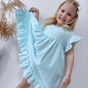 Puede incluir: Un vestido azul claro hecho de tela de muselina texturizada con mangas cortas con volantes, cuello redondo y un dobladillo ancho con volantes que se extiende a lo largo del lateral. La prenda tiene un corpiño suelto y fruncido.