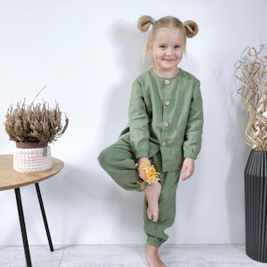 Puede incluir: Un conjunto de camisa de manga larga abotonada y pantalones con pu&ntilde;os el&aacute;sticos en color verde salvia. La camisa presenta un cuello redondo y cuatro botones de madera clara en la parte delantera. La tela tiene un tejido texturizado visible.