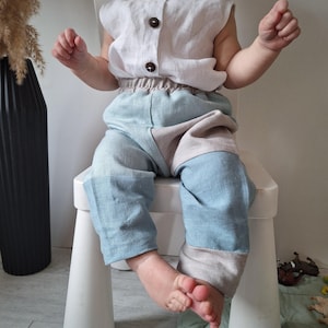 Op de afbeelding: Een baby-outfit met een mouwloze top met knopen en een broek met kleurblokken. De top is lichtbeige en de broek combineert lichtblauwe en beige panelen. De outfit is gemaakt van linnenachtige stof, geschikt voor een peuter.