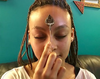 May include: A person with braided hair holds a silver incense stick with a decorative lotus flower design in front of their forehead. The incense stick is emitting smoke.
